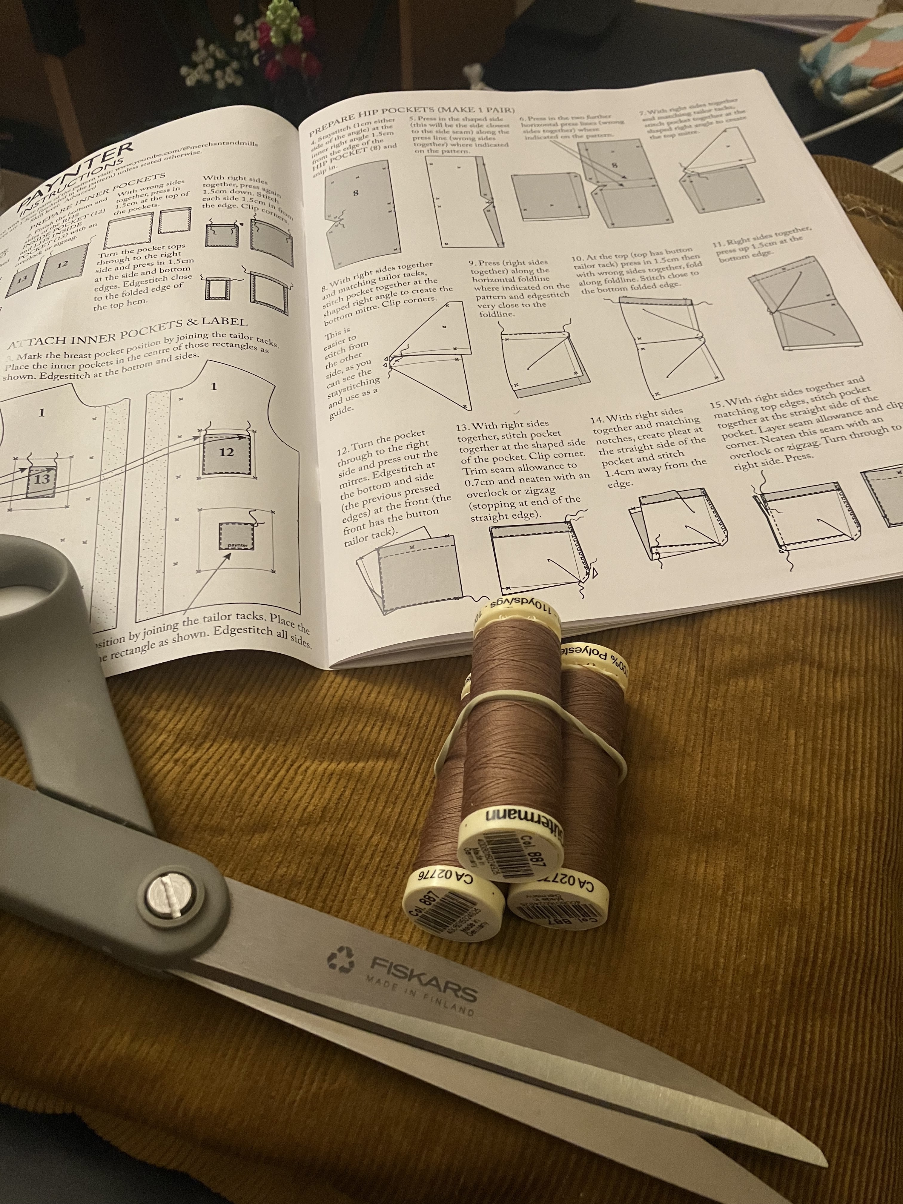 Photo d’ambiance avec du tissu en velours côtelé, trois bobines de fil de même couleur, une paire de ciseaux à tissu, et la notice d’assemblager d’une veste.