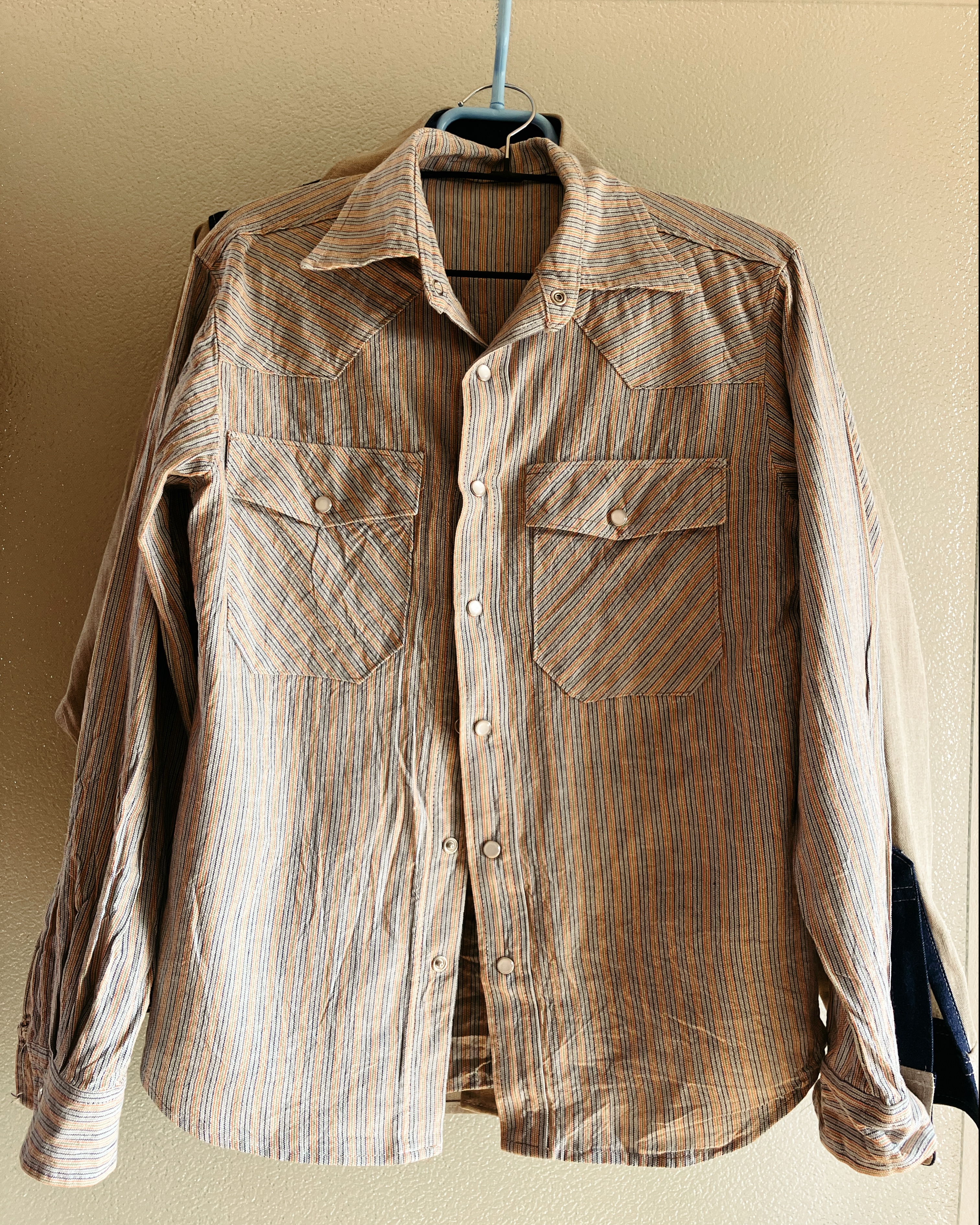 Photo d’une chemise qui pend à un cintre, elle est blanche avec des rayures fines et des boutons pression nacrés.