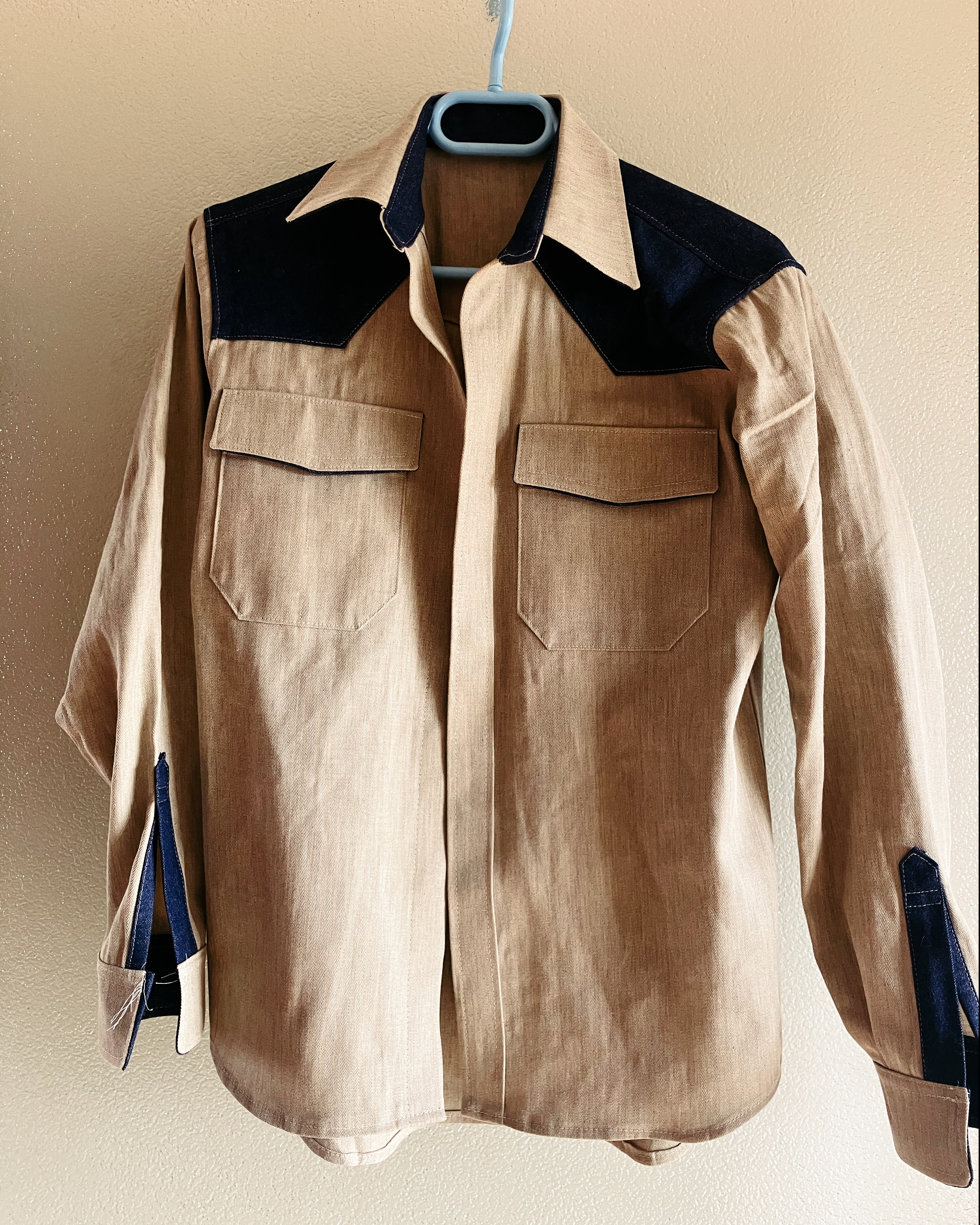 Photo d’une chemise qui pend à un cintre. Elle est beige, avec les épaules bleues jeans.