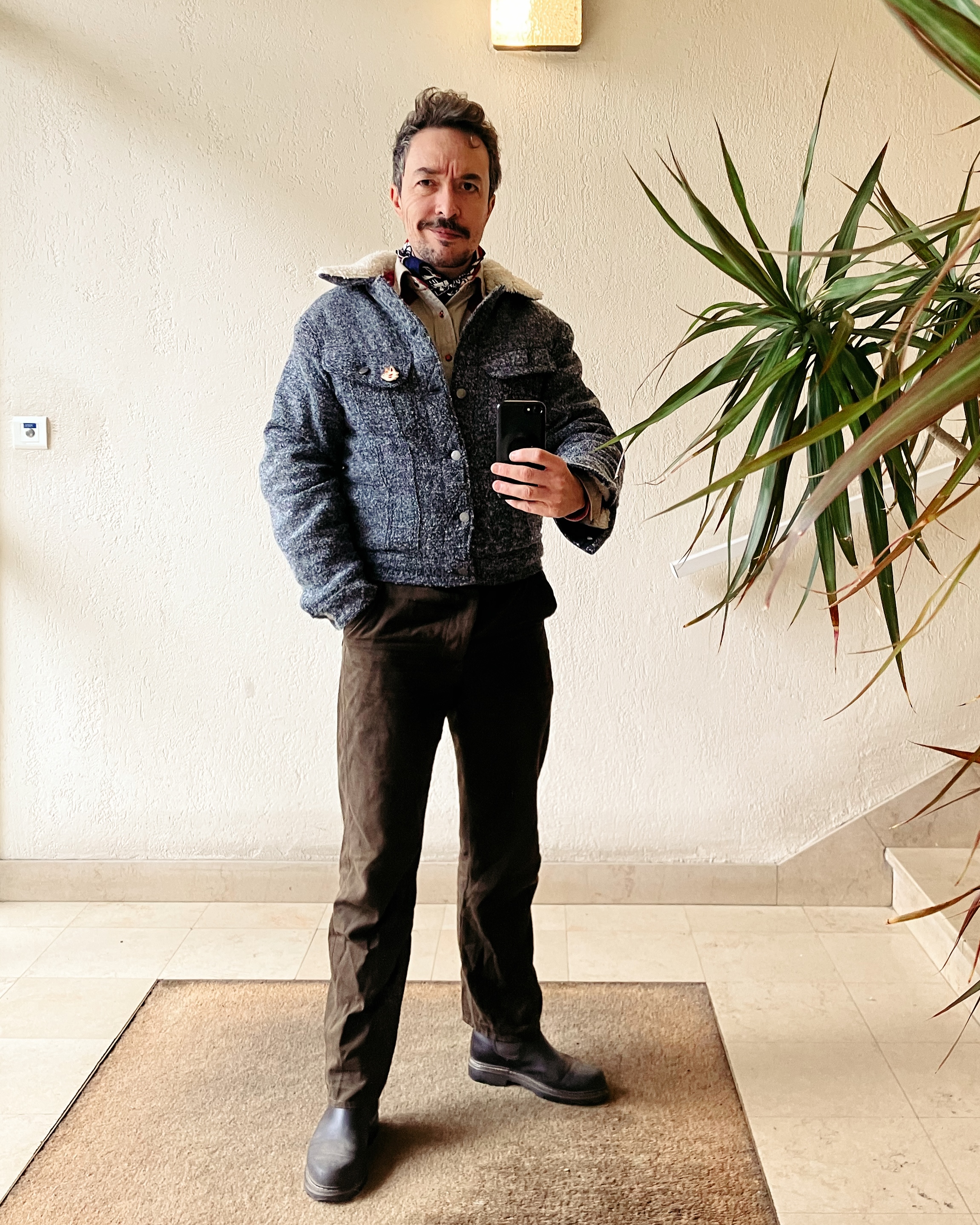 Photo d’un homme moustachu devant un miroir. Il porte un blouson en tissu chiné gris avec un col en fausse fourrure type sherpa, un pantalon fin brun/vert.