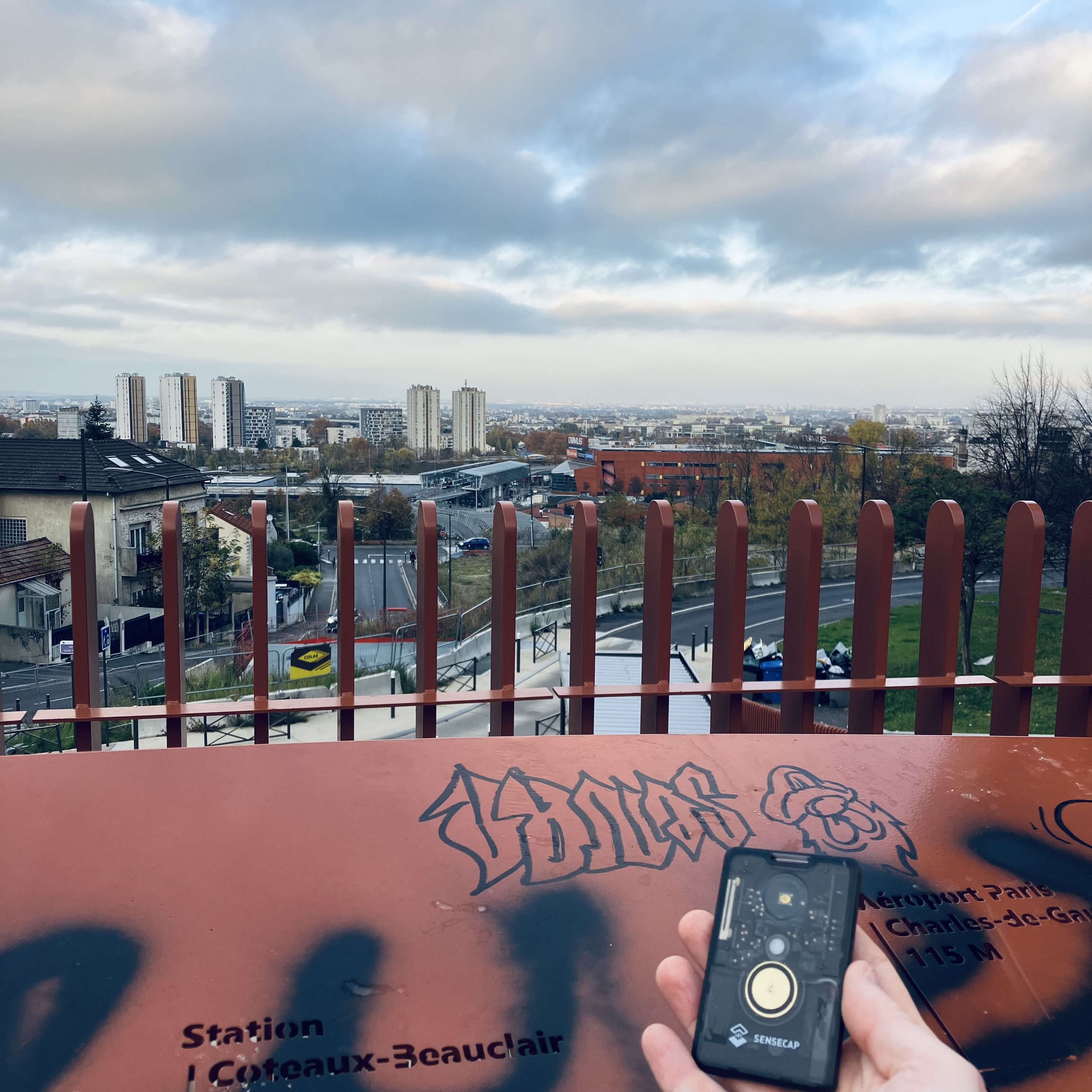 Photo d'un paysage de ville pris depuis un point de vue en hauteur. Au premier plan, une main tient un petit gadget en plastique noir translucide de la taille d'une carte bleue.