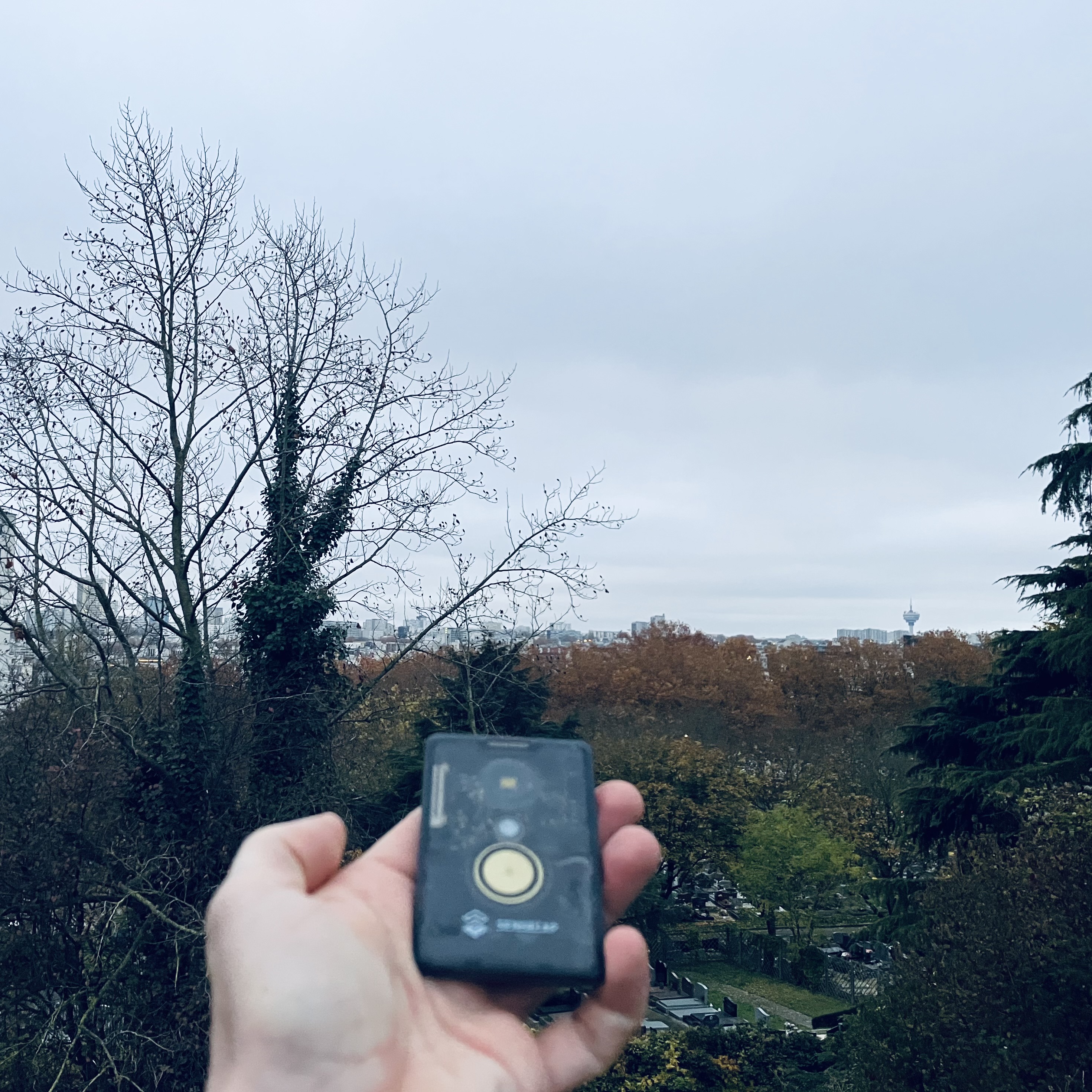 Photo d'un paysage de ville pris depuis un point de vue en hauteur, on voit la tour de Romainville. Au premier plan, une main tient un petit gadget en plastique noir translucide de la taille d'une carte bleue.