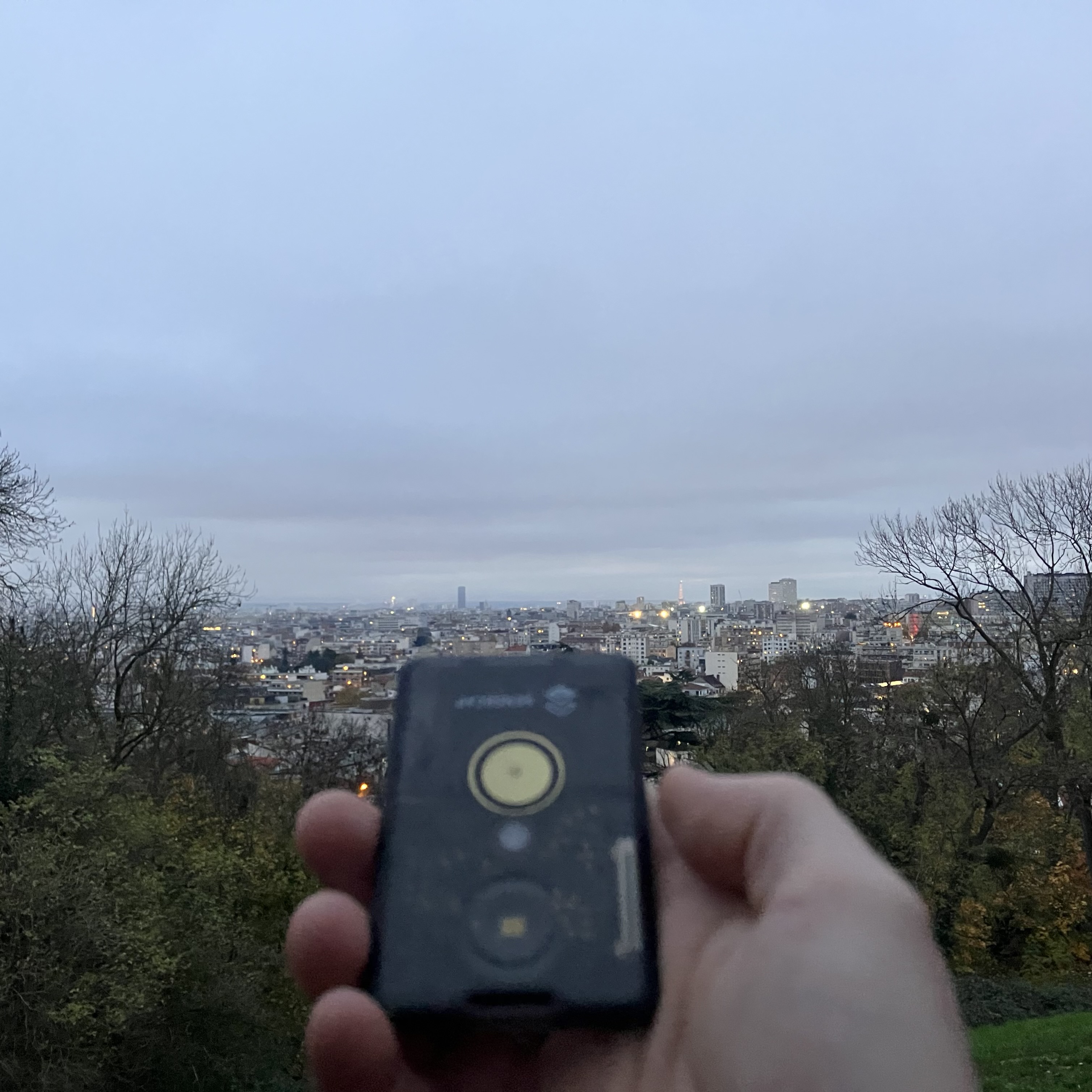 Photo d'un paysage de ville pris depuis un point de vue en hauteur, on peut voir en tout petit la tour Eiffel. Au premier plan, une main tient un petit gadget en plastique noir translucide de la taille d'une carte bleue.