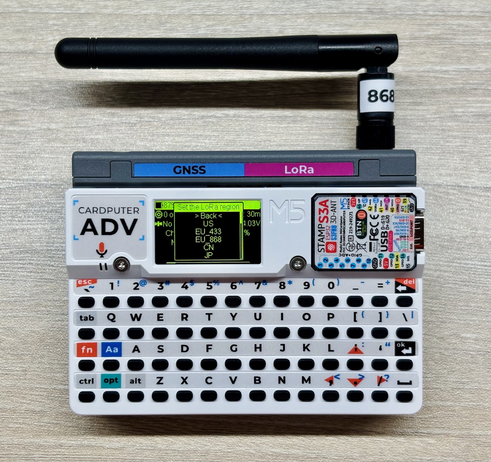 Une sorte de mini-micro-ordinateur composé d’un clavier de tout petits boutons sur un boitier rectangulaire gris, un petit écran noir à écritures vertes, et au sommet un module gris d’où s’échappe une antenne. Le tout donne une impression de jouet mais technologique.