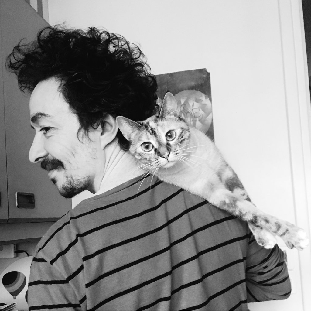 Portrait en noir et blanc d’un homme avec une moustache, des cheveux bouclés et un haut à rayures, avec son chat mignon sur l’épaule.